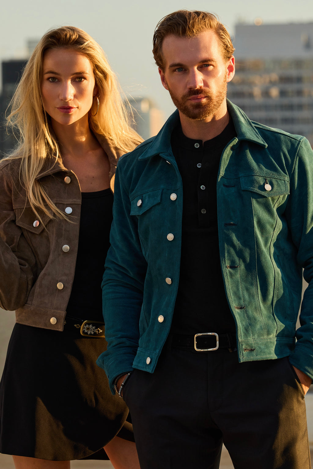 A man and a woman standing outdoors at sunset wearing suede jackets. The man wears a teal-green jacket over a black henley shirt and dark pants, while the woman wears a brown jacket over a black top and skirt.