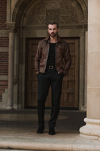 Man wearing a brown leather jacket standing in front of an ornate door.