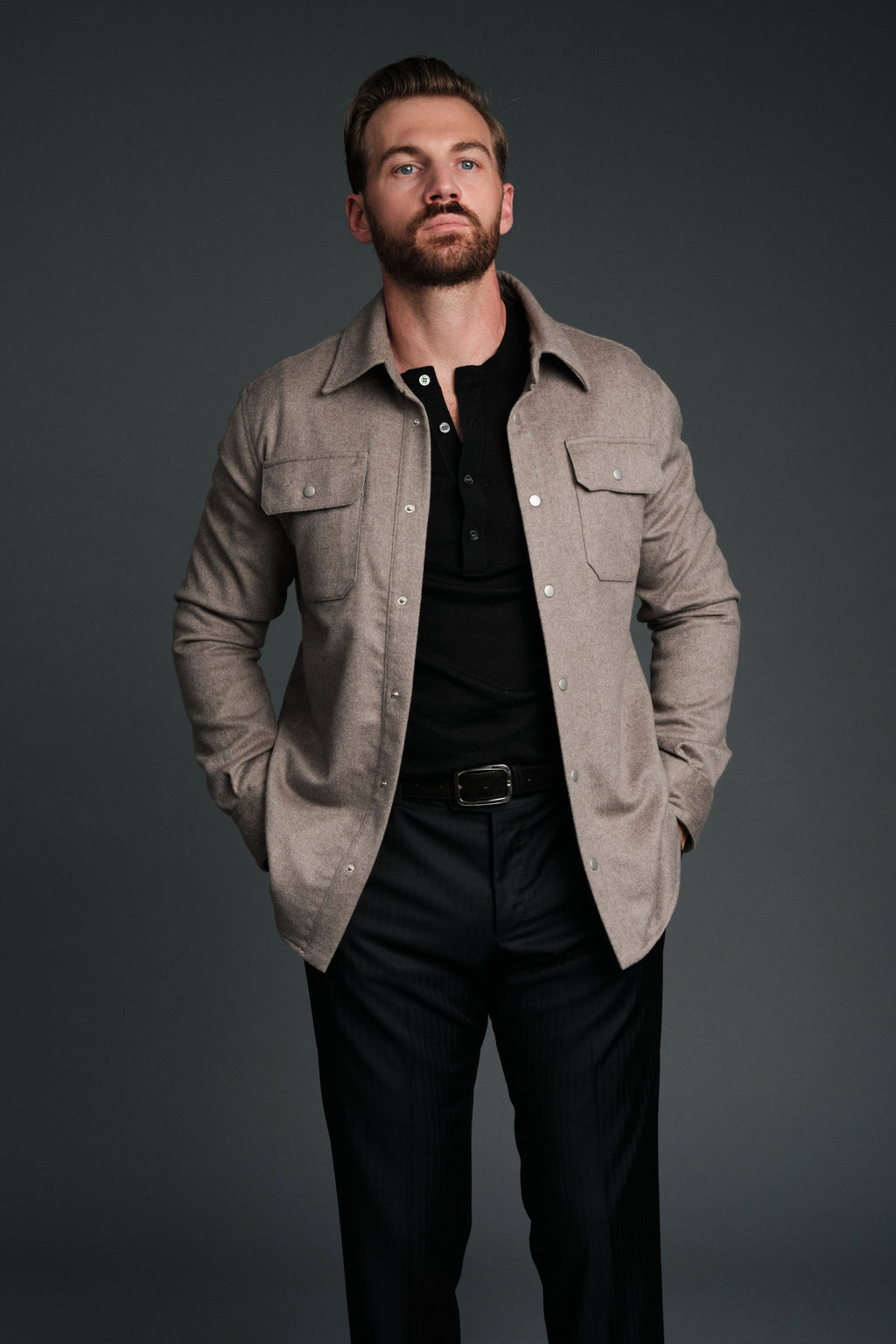 Man wearing a beige jacket over a black shirt and pants on a dark background