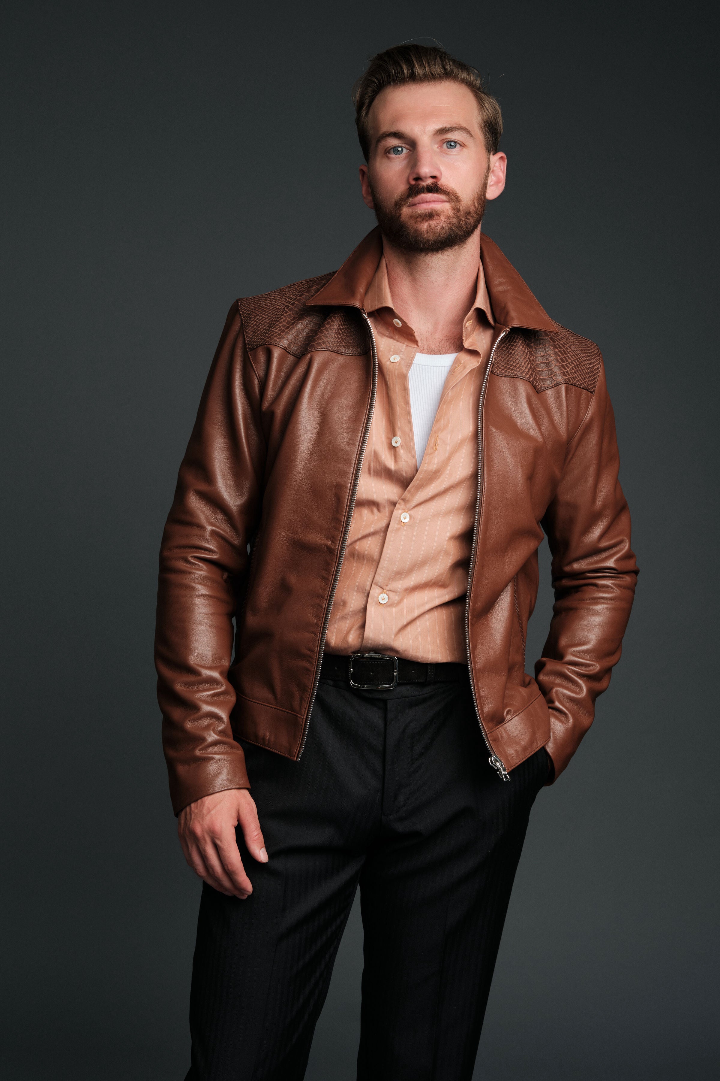Man wearing a brown leather jacket over a white shirt on a dark background
