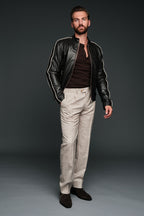 Man wearing a black brown leather jacket, brown shirt, and beige pants against a dark background