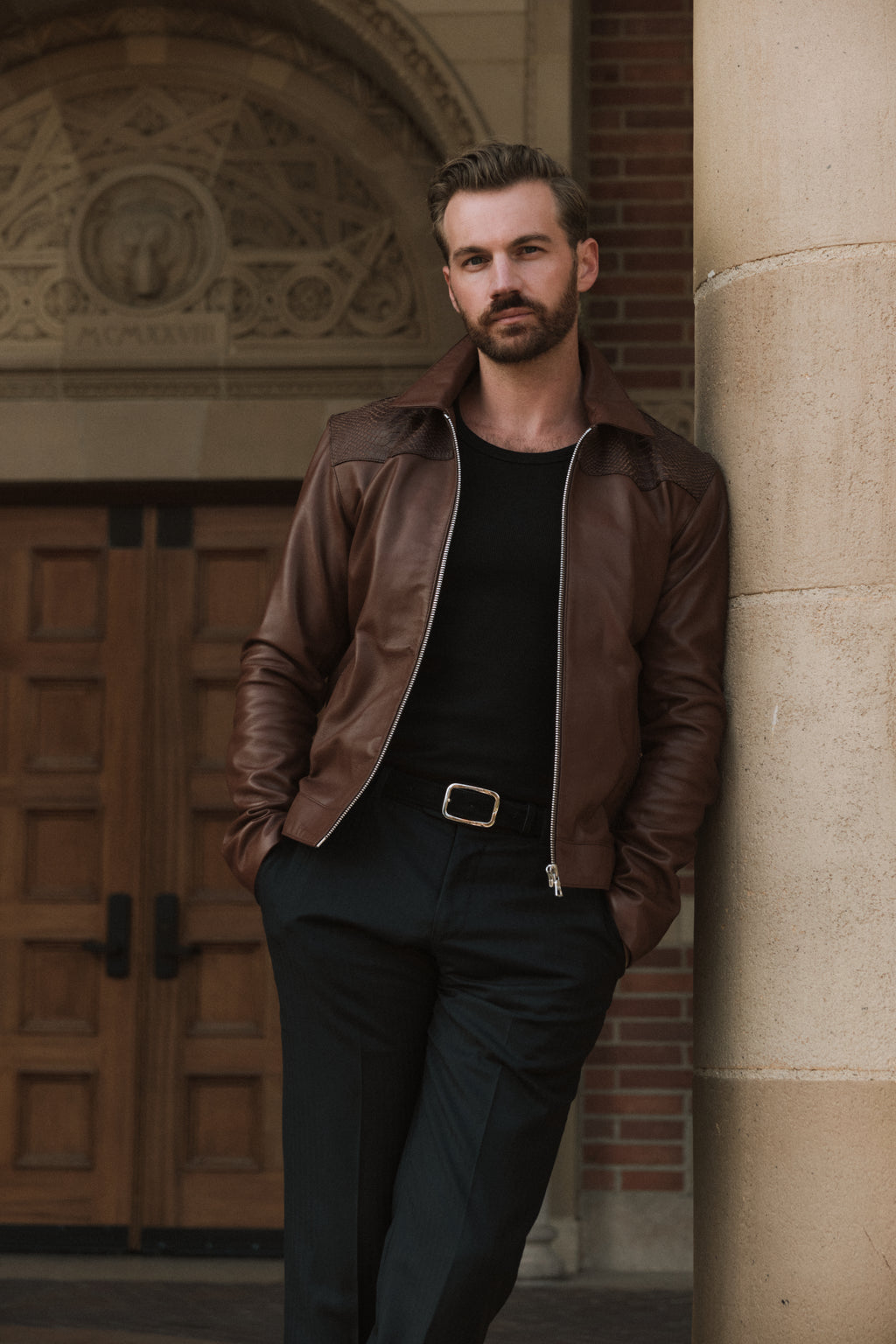 Man in a brown leather jacket and black outfit leaning against a stone wall.
