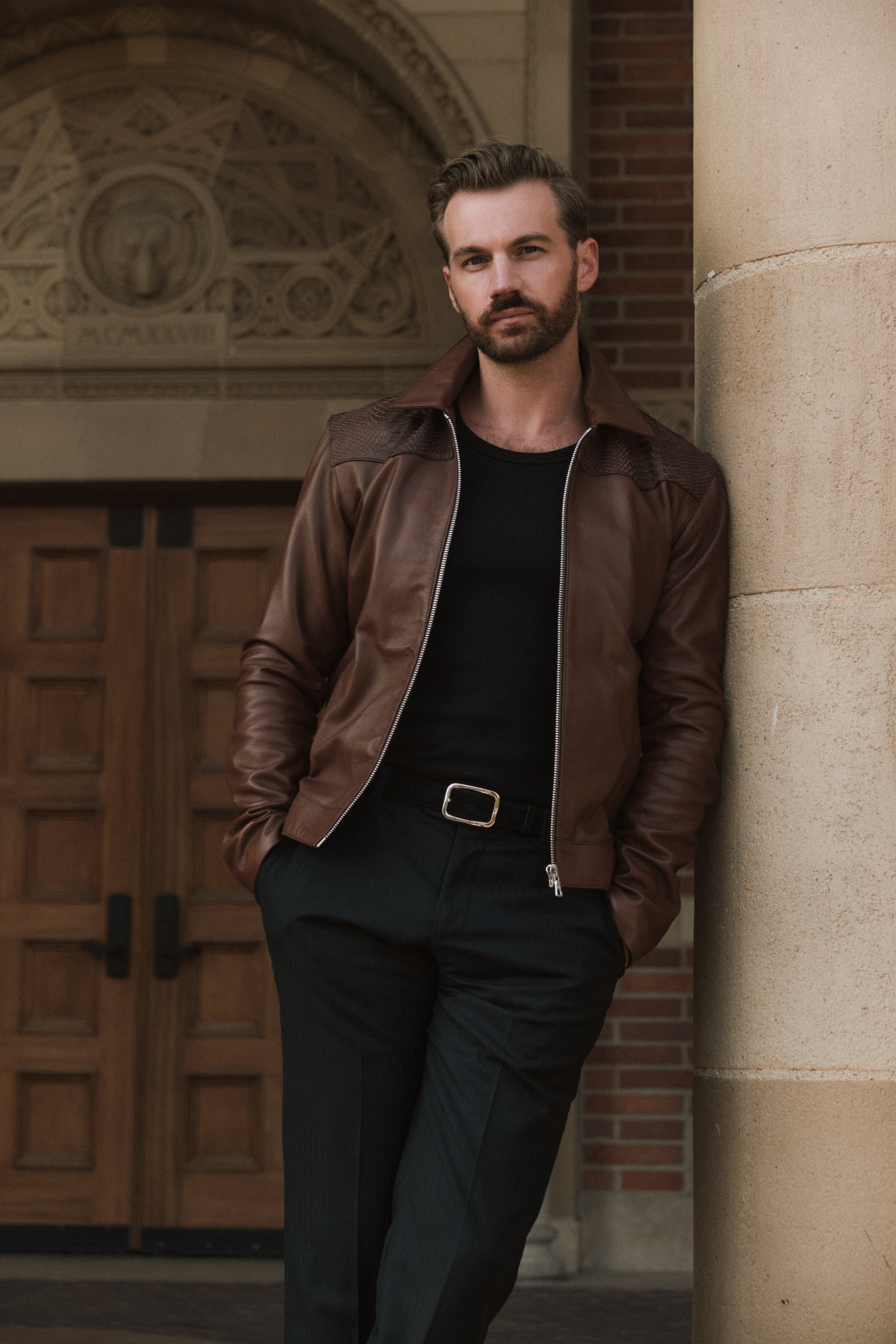 Man in a brown leather jacket and black outfit leaning against a stone wall.