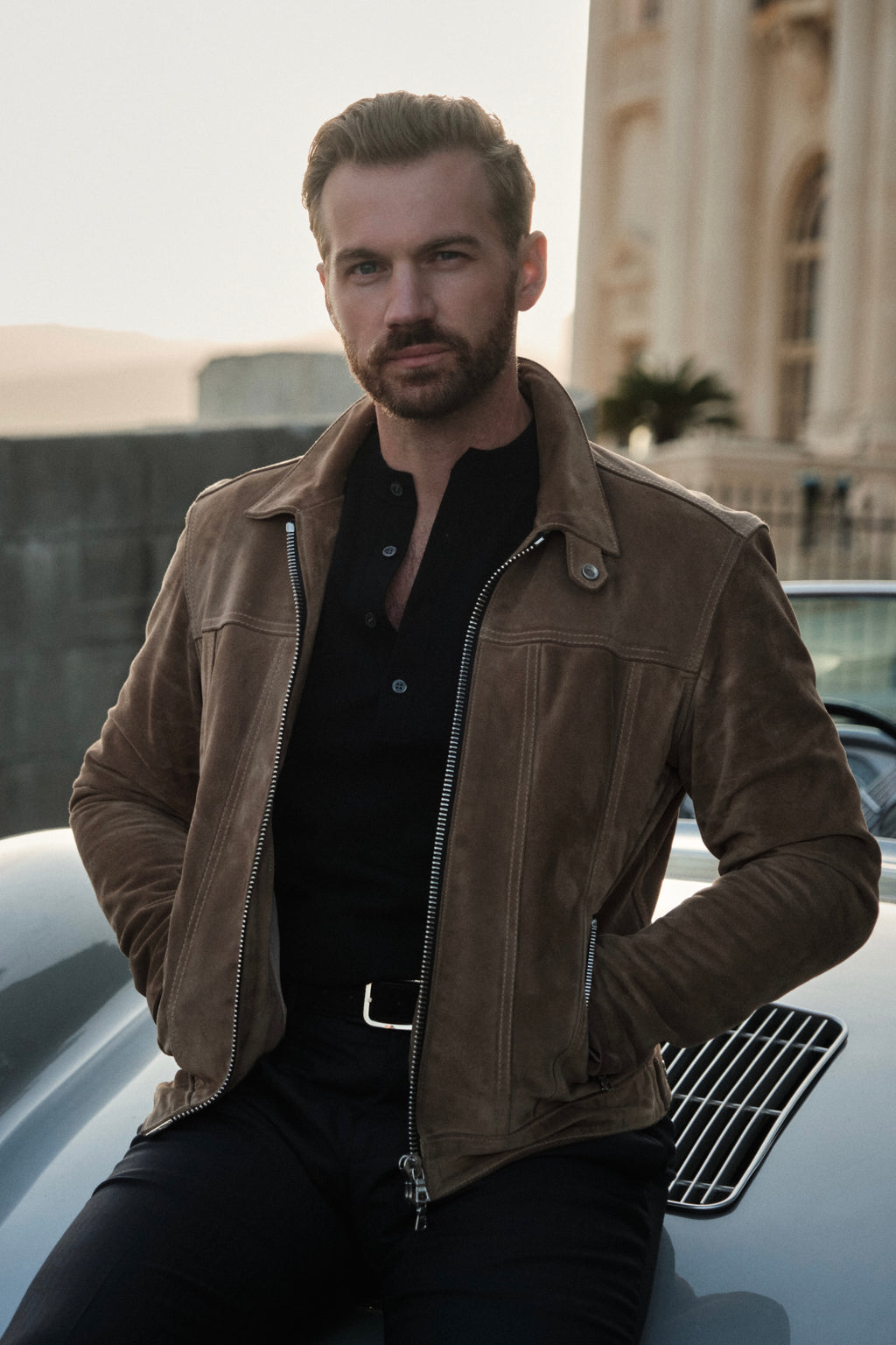 Man in a brown leather jacket sitting on a car with a building in the background