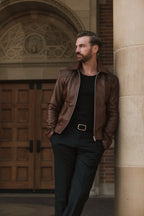 Man in a brown leather jacket leaning against a stone column with wooden doors in the background