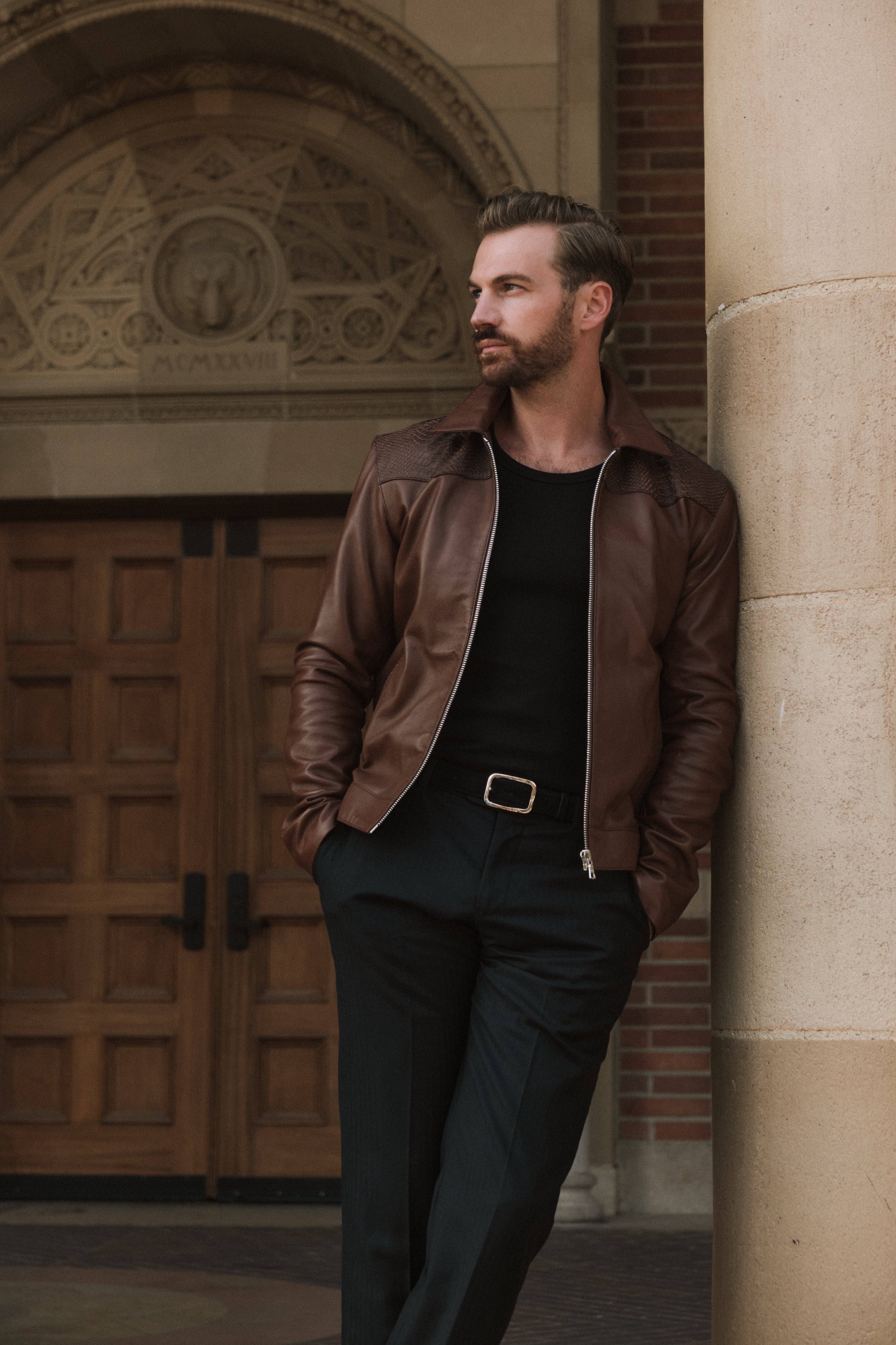 Man in a brown leather jacket leaning against a stone column with wooden doors in the background