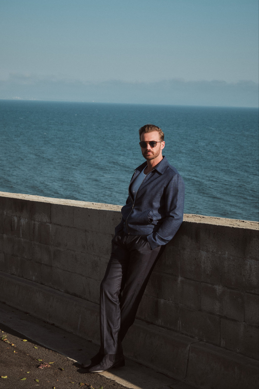 Man in a blue shirt and sunglasses leaning against a wall with ocean in the background