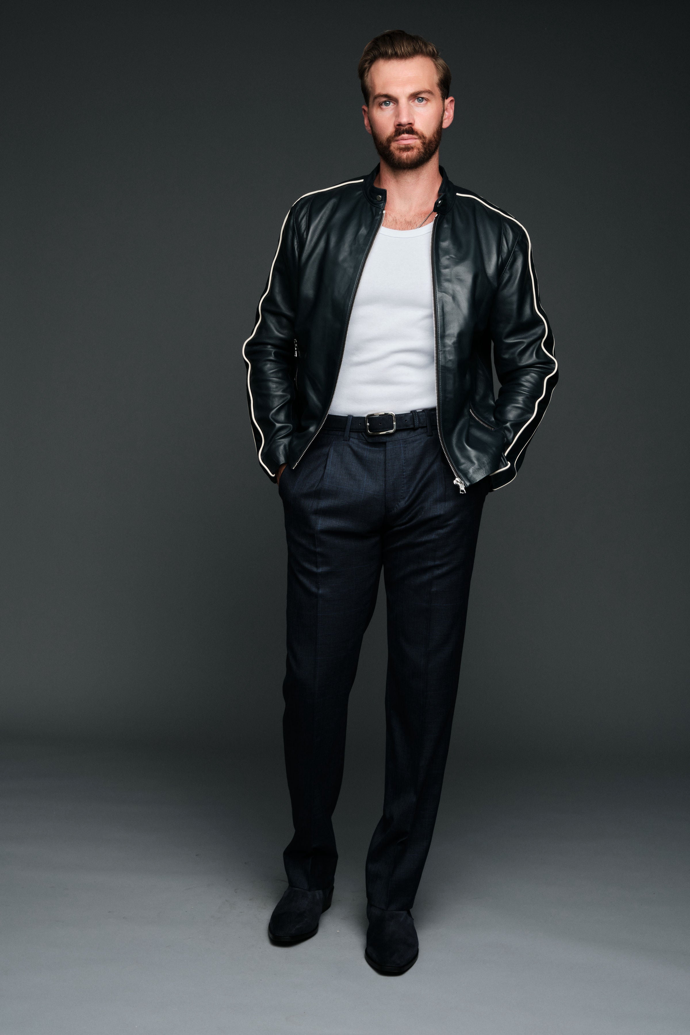 Man wearing a navy leather jacket with white stripes, white shirt, and dark pants on a gray background