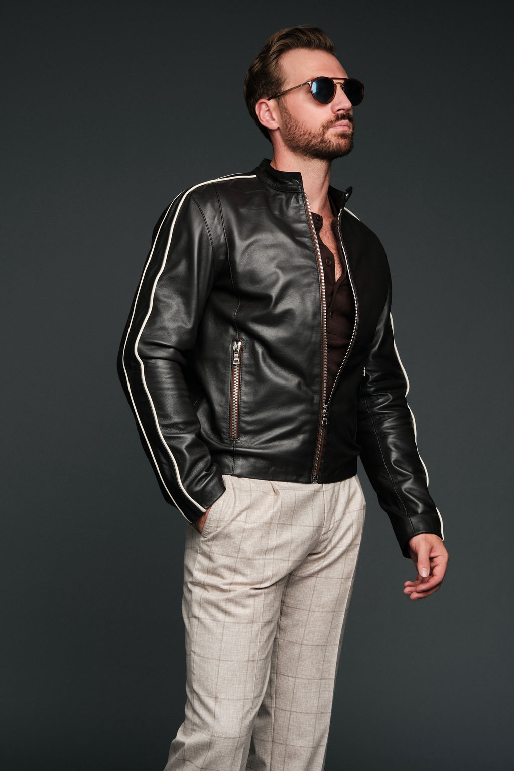 Man wearing a black brown leather jacket with white stripes and sunglasses on a dark background