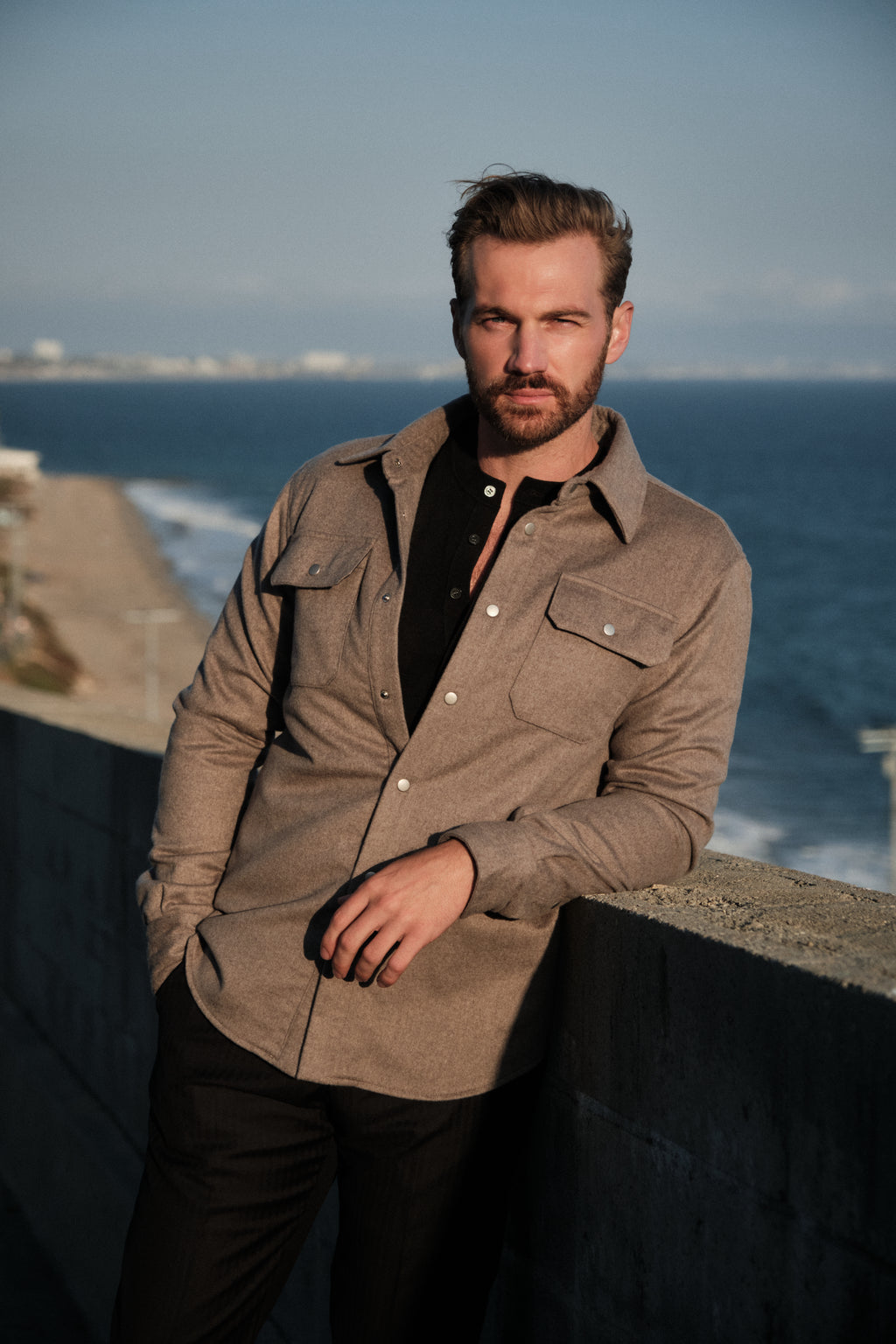 Man standing by the ocean wearing a brown jacket and black shirt.