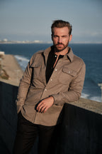 Man standing by the ocean wearing a brown jacket and black shirt.