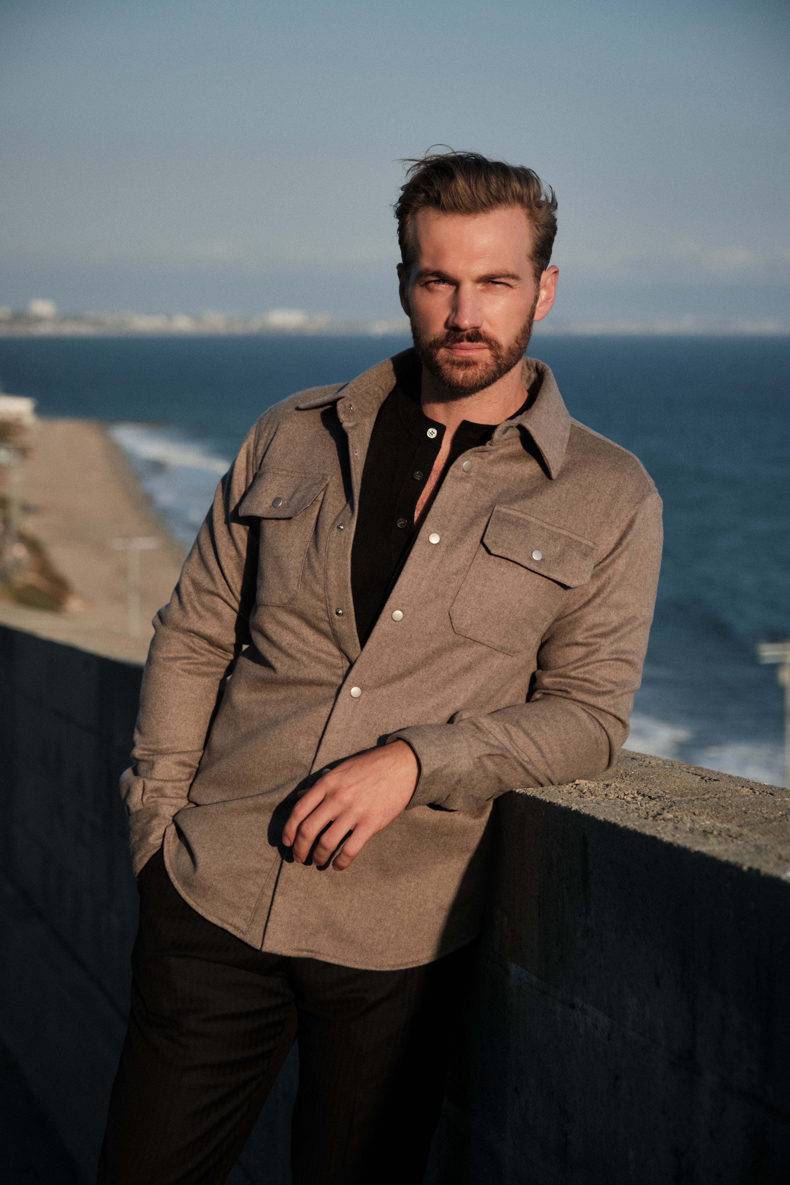 Man standing by the ocean wearing a brown jacket and black shirt.