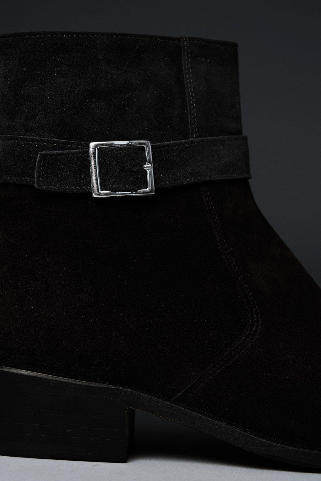 Black suede ankle boot with a silver buckle on a dark background