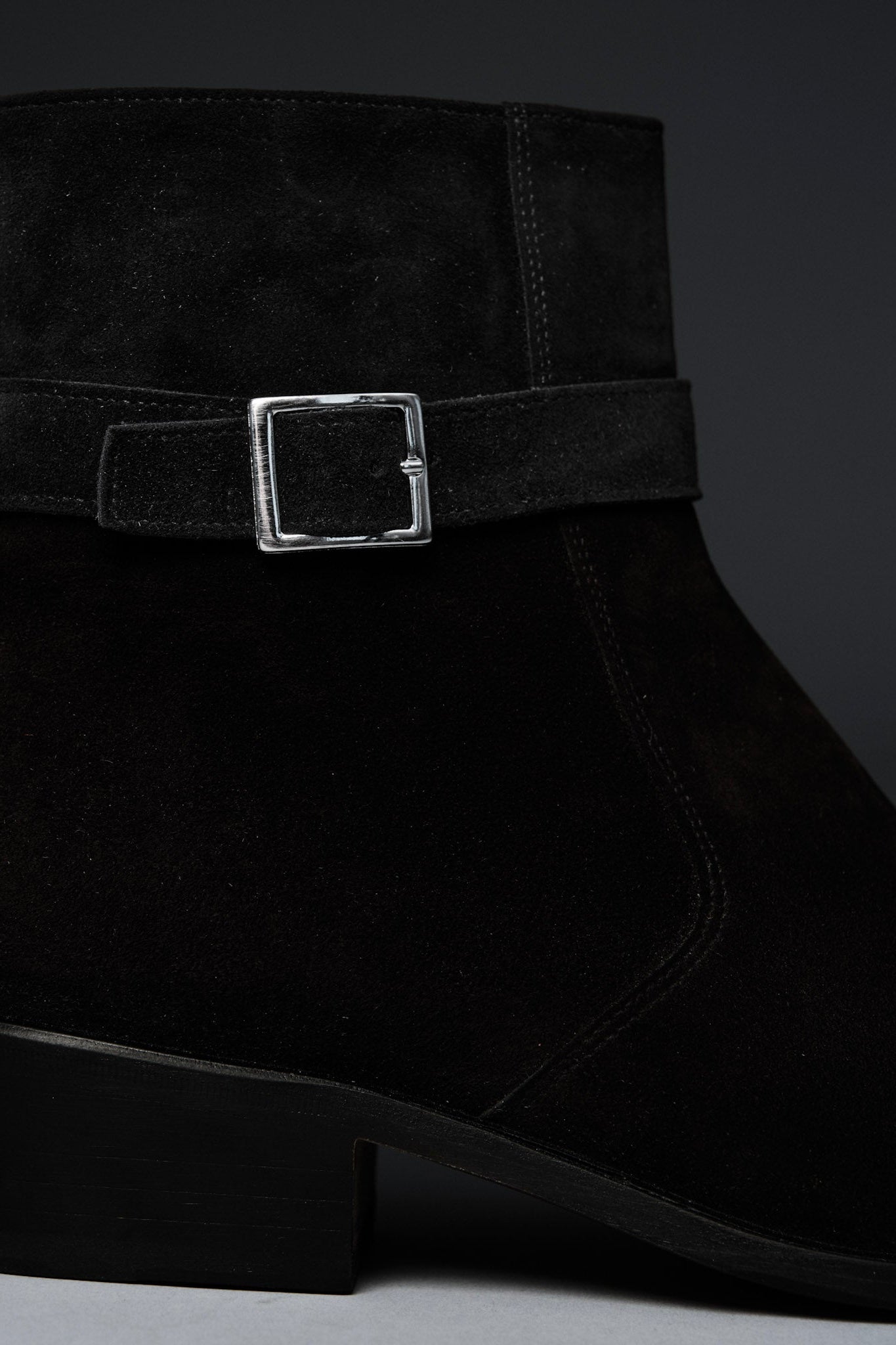 Black suede ankle boot with a silver buckle on a dark background